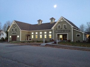 An outdoor, wide-angle shot of Tamworth Distilling & Mercantile, a large, olive-green shingle-sided building with white trim. The building features two prominent gables with decorative white timber framing, a dark gray shingled roof, and two small cupolas.