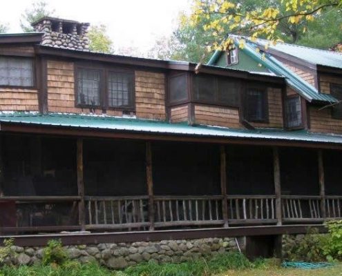 An outdoor, eye-level shot of a large, rustic two-story cabin with a mix of cedar shingle siding and dark brown trim. The most prominent feature is a long, screened-in porch with a rustic log railing that spans the entire front of the building. The cabin features a green metal roof with several gabled sections and a prominent stone chimney on the left.