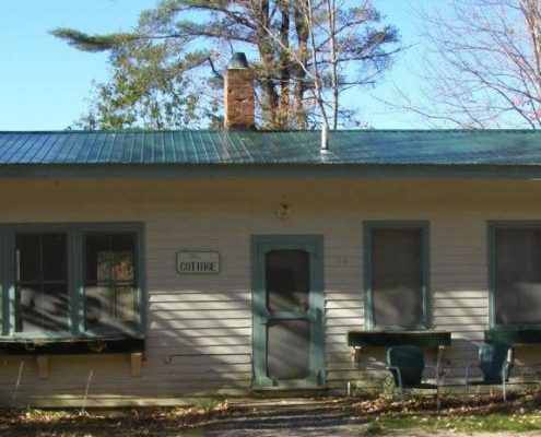An outdoor, eye-level shot of a single-story cottage with beige horizontal siding and a dark green metal roof. The cottage features a central screen door flanked by several multi-pane windows with dark green trim and matching wooden window boxes.
