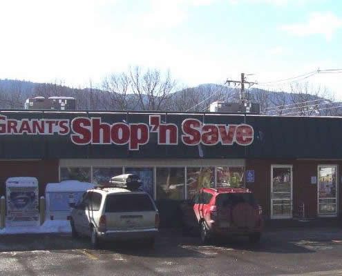 An outdoor, eye-level shot of Grant's Shop 'n Save, a single-story grocery store with reddish-brown siding and a long, dark green metal roof. A large red and white sign with the store's name is mounted on the roofline above a large storefront window.