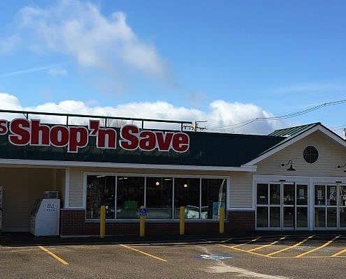 An outdoor, wide-angle shot of Grant's Shop 'n Save, a single-story grocery store with cream-colored horizontal lap siding and a dark green gabled roof. A large sign with red and white lettering identifies the store above a recessed entrance area with automatic glass sliding doors.