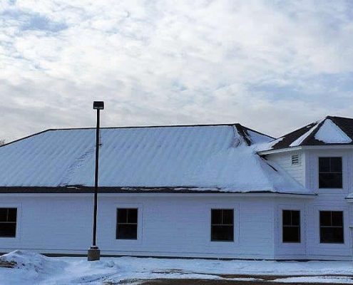 An outdoor, wide-angle shot of the North Conway Water Precinct Office, a single-story white building with horizontal lap siding and several gabled roof sections. The scene is set in winter, with a light dusting of snow on the gray shingled roofs and large piles of plowed snow along the building's base.