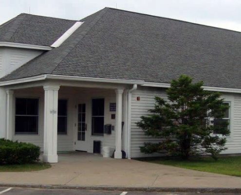 An outdoor, eye-level shot of a single-story white horizontal-lap sided office building with a dark gray shingled gabled roof. The building features several white-trimmed double-hung windows and a central, recessed entrance area supported by a thick white column.