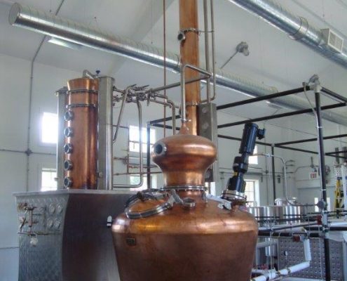 An indoor, eye-level shot of a large, gleaming copper distilling apparatus in a modern industrial space. The central focus is a bulbous pot still on four legs, connected by a network of stainless steel pipes to a tall, vertical column still with multiple circular sight glasses.