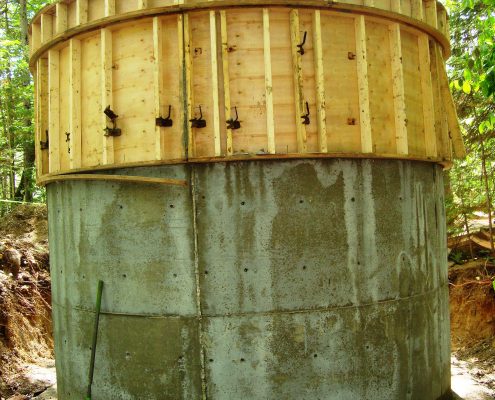 An outdoor, eye-level shot of a large, cylindrical concrete structure under construction with the bottom half made of smooth, gray poured concrete with visible horizontal seams and small, evenly spaced holes. A large wooden circular form sits on top of the concrete, featuring numerous vertical wood studs.