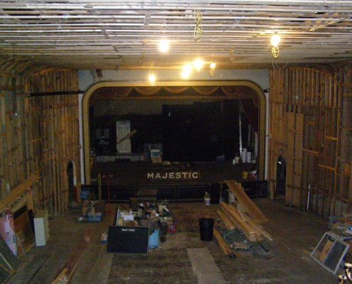 The walls and curved ceiling of the Majestic Theater are stripped to their exposed wooden framing, with some of the original blue plaster still visible at the top. The center of the room is cluttered with construction debris.