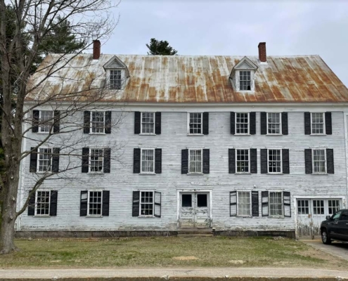 The North Conway House or Bunker Building is a large, three-story wood-frame building with a weathered white facade and black shutters. A steep pitched metal roof is showing rust and wear.
